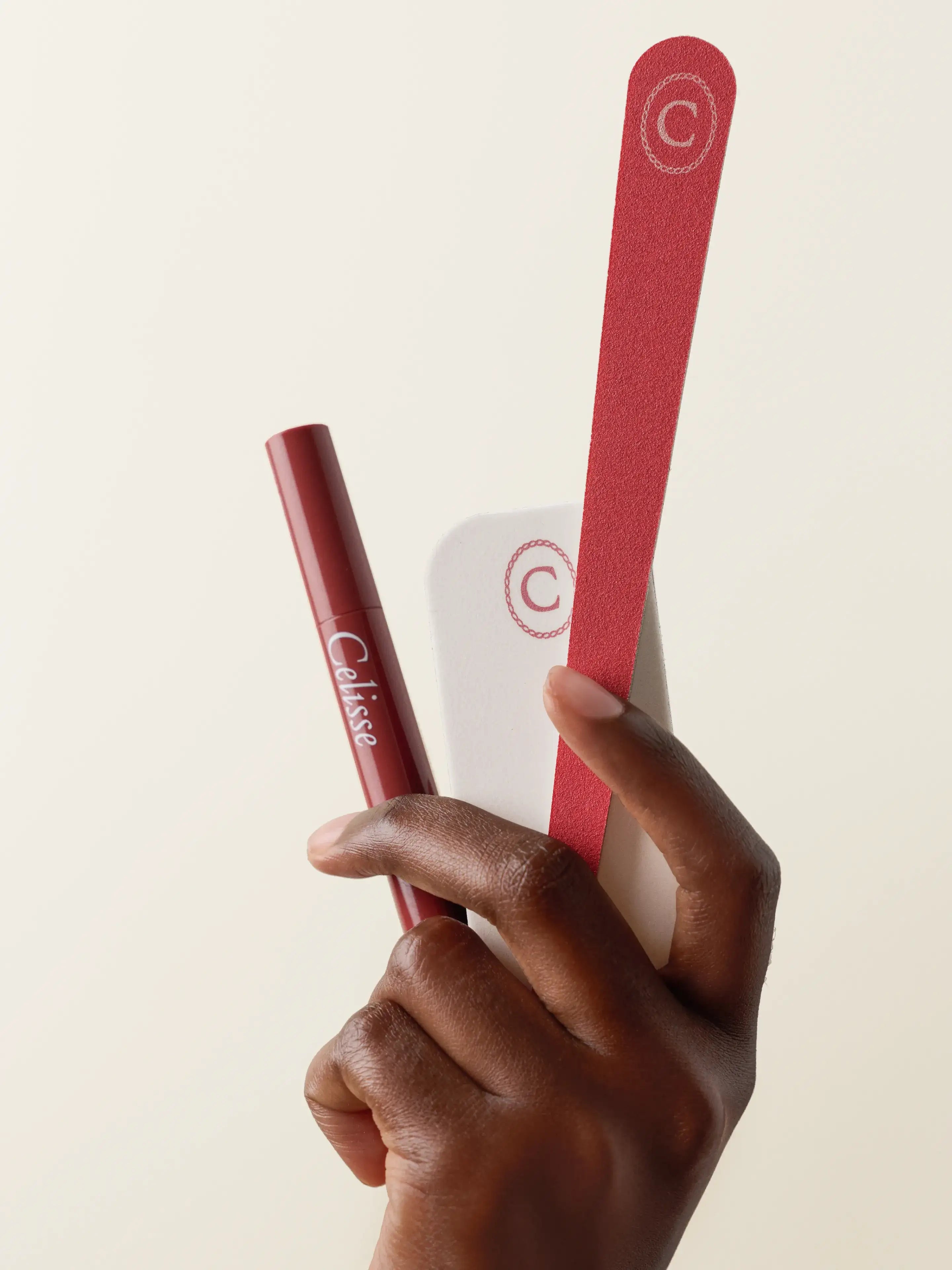 Hand holding a red nail file, white buffer, and dark red cuticle serum pen.